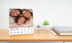 Tischkalender mit Familie auf Tisch, daneben Wecker und Pflanze